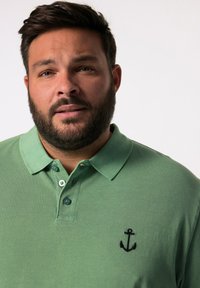 Grön bomullspolo med en treknapps placket, broderad svart ankarl logo på vänster bröst och en texturerad tygoverflöde.