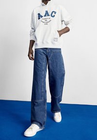 Grå hoodie med "AAC"-logotyp och känguruficka, kombinerad med vida blå jeans och vita sneakers, framför en blå golvbakgrund.