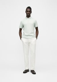 Homme debout, mains dans les poches, portant un polo vert clair à manches courtes, un pantalon blanc et des mocassins marron foncé sur fond blanc.