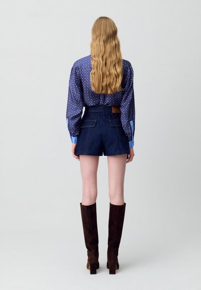Camisa azul de lunares con mangas largas, combinada con pantalones cortos de mezclilla oscuros que tienen bolsillos traseros y un parche de cuero marrón. Botas marrones hasta la rodilla.