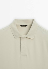 Polo en coton beige clair avec un col et une courte patte de boutonnage, comportant une petite étiquette de marque noire à l'intérieur du col.