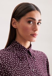 Bordeaux geknoopte blouse met witte stippen, voorzien van een klassieke kraag en een textuurstof, acentuated door paarse knopen.