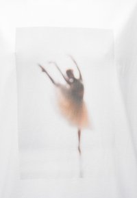 Biała koszulka z rozmytym nadrukiem tancerki baletowej w ruchu, z delikatnym gradientem od ciemnych do jasnych tonów, bez wyraźnych detali.