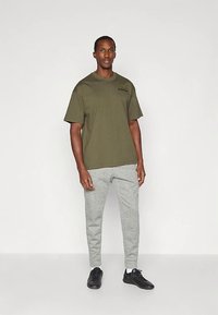 Vīrietis stāv, ģērbies olīvzaļā pārāk lielā T-kreklā, gaiši pelēkās sporta biksēs, baltās zeķēs un melnās sporta apavās uz baltas fona.