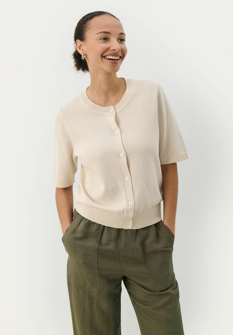 Cardigan beige à manches courtes avec col rond et fermeture à boutons, confectionné en tissu doux. Présente un ourlet et des poignets côtelés. Associé à un pantalon vert olive.