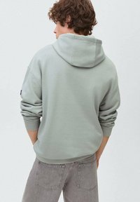 Ljusgrå huvtröja med ribbade muddar och fåll, med en avslappnad passform. Buren med grå byxor, sedd bakifrån.