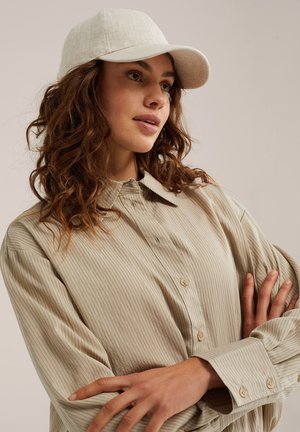 Ung kvinde med krøllet brunt hår iført en beige skjorte med knapper og lysebeige baseballkasket, armene krydset.