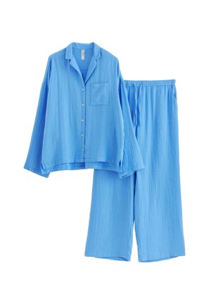Completo pigiama azzurro chiaro realizzato in tessuto testurizzato. Include una camicia button-up rilassata con tasca sul petto e pantaloni a gamba larga con vita elasticizzata.