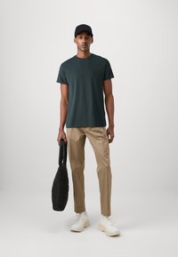 Dunkelgrünes Rundhals-T-Shirt, hellbraune Hose, weiße Sneakers und eine schwarze, strukturierte Tote-Tasche. Einfaches, lässiges Outfit ohne Muster.