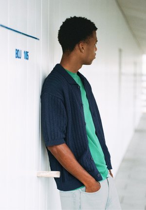 Jeune homme aux cheveux courts et bouclés appuyé contre un mur blanc, portant un cardigan bleu foncé à manches courtes par-dessus une chemise verte et un pantalon clair.