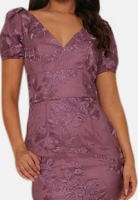 Laventelinvärinen mekko, jossa V-aukko, lyhyet pörröiset hihat ja kukkaistikkaus. Istuvamallinen design, jossa sileä kangaspinta.