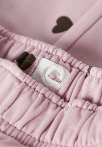 Tejido de color rosa claro con textura arrugada, con una etiqueta blanca y un botón transparente. Patrón de corazones marrones visible en el fondo.