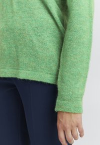 Grön, stickad tröja med en mjuk, fluffig textur. Har en ribbad mudd och en något kortad avslutning. Buren över marinblå byxor.