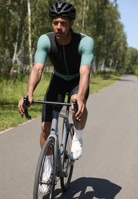 Cyklist på en grå väg iklädd en svart och turkos cykeltröja samt svarta shorts, som cyklar på en silverfärgad landsvägscykel med nedåtriktade styren.