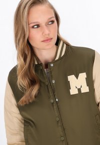 Giacca bomber verde e beige con colletto lavorato a maglia, bottoni a pressione e una grande lettera "M" color crema sul lato sinistro del petto. Tessuto dalla texture liscia.