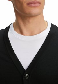 Cardigan nero con design a scollo a V, texture lavorata a maglia, con chiusura a un bottone. Indossato sopra una maglietta bianca.