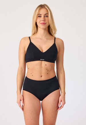 Femme aux cheveux blonds portant un bralette noir et un sous-vêtement taille haute, debout devant un fond blanc uni.