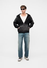 Νέος άνδρας που φοράει μαύρο φούτερ με φερμουάρ, λευκό πουκάμισο, φαρδιά μπλε τζιν, μαύρα γυαλιά ηλίου και αθλητικά παπούτσια, στέκεται με τα χέρια στις τσέπες.