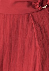 Červená, texturovaná tkanina s nařaseným pasem a splývavým designem. Hladký povrch s jemnými vráskami, které zvýrazňují strukturu oděvu.