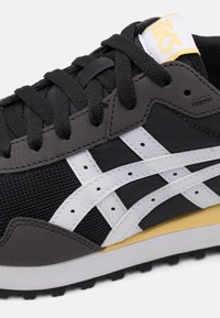 Black and gray sneakers with white accents, textured mesh and leather materials, featuring black laces and a yellow logo on the tongue.