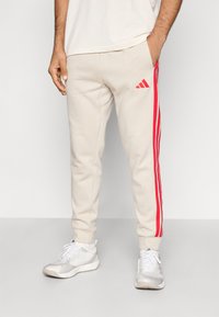 Jasnobeżowe spodnie dresowe z czerwonymi bocznymi paskami, elastyczną talią i ściągaczami na nogawkach. Z przodu małe czerwone logo Adidas.