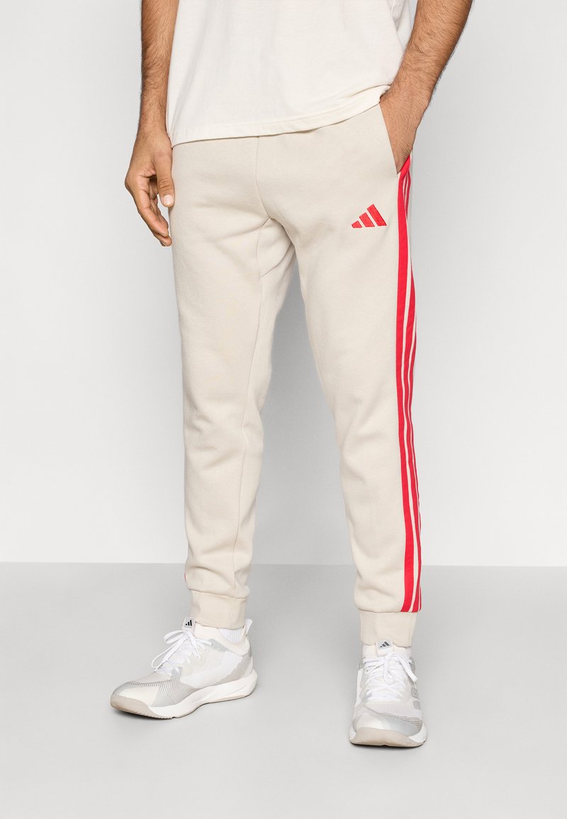 Jasnobeżowe spodnie dresowe z czerwonymi bocznymi paskami, elastyczną talią i ściągaczami na nogawkach. Z przodu małe czerwone logo Adidas.