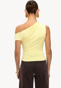Haut jaune ajusté à une épaule en tissu doux avec des détails froncés, associé à un pantalon large marron foncé. Des accessoires marron complètent le look.