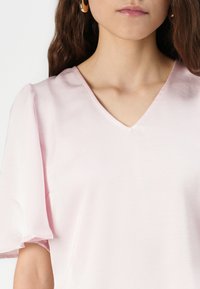 Blusa de color rosa claro con escote en V y mangas cortas abullonadas. La tela tiene una textura suave y detalles de costura mínimos cerca del escote.