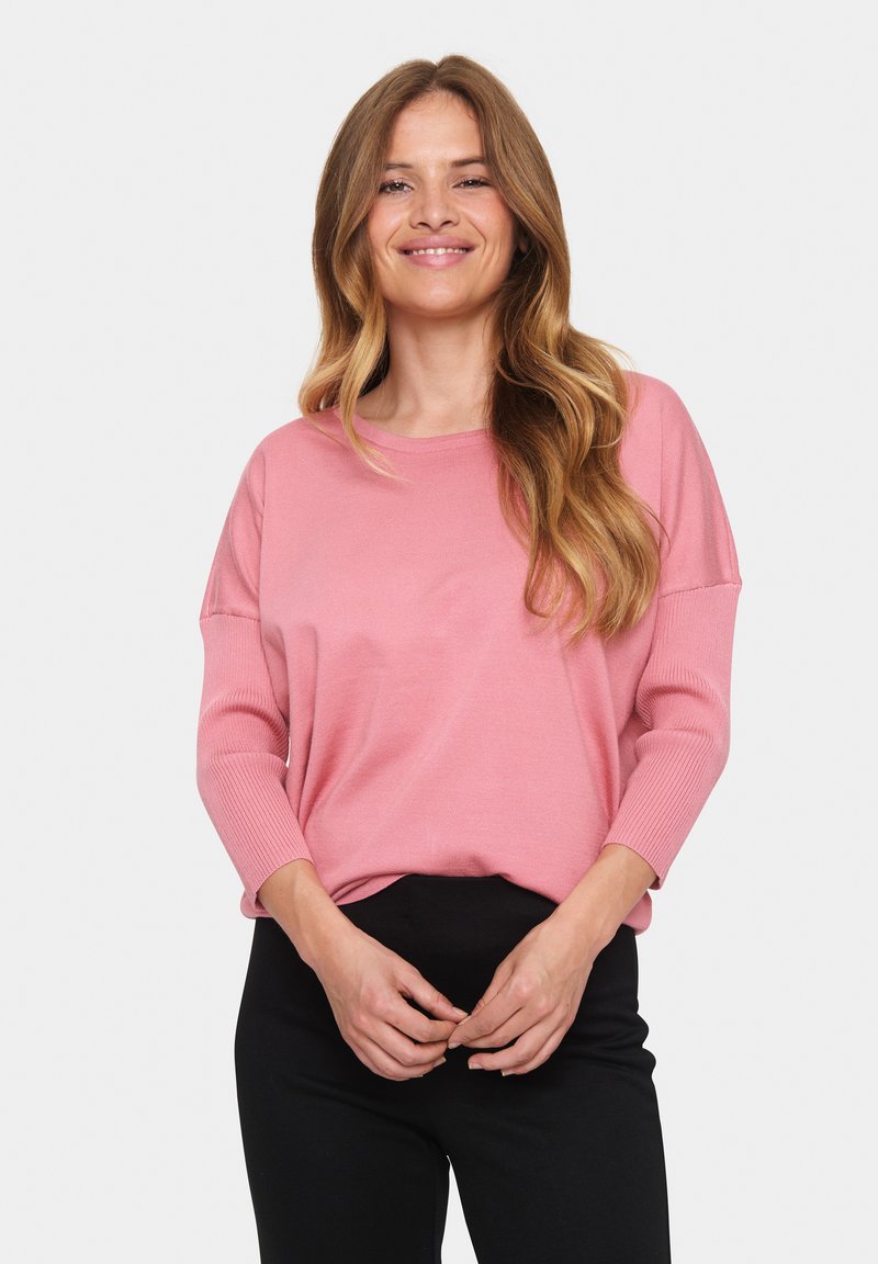 Rosa Strickjacke mit 3/4-Ärmeln, Rundhalsausschnitt und gerippter Textur, kombiniert mit schwarzen Hosen. Weite Passform, glattes Material.