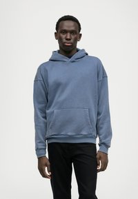 Hoodie azul em algodão com um bolso frontal, capuz com cordão e punhos canelados, combinado com calças pretas. Textura suave e ajuste descontraído.