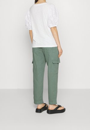 Femme portant un chemisier blanc à manches bouffantes, un pantalon cargo vert et des sandales compensées noires, debout devant un fond uni.