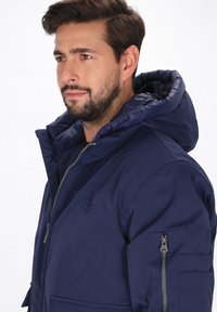 Sötétkék kapucnis dzseki sima textúrával, elöl cipzárral, oldalsó zsebekkel és mellkasi logóval.