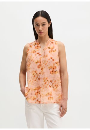 Femme aux longs cheveux foncés portant un chemisier sans manches pêche avec un motif floral orange et un pantalon blanc, se tenant devant un fond blanc.