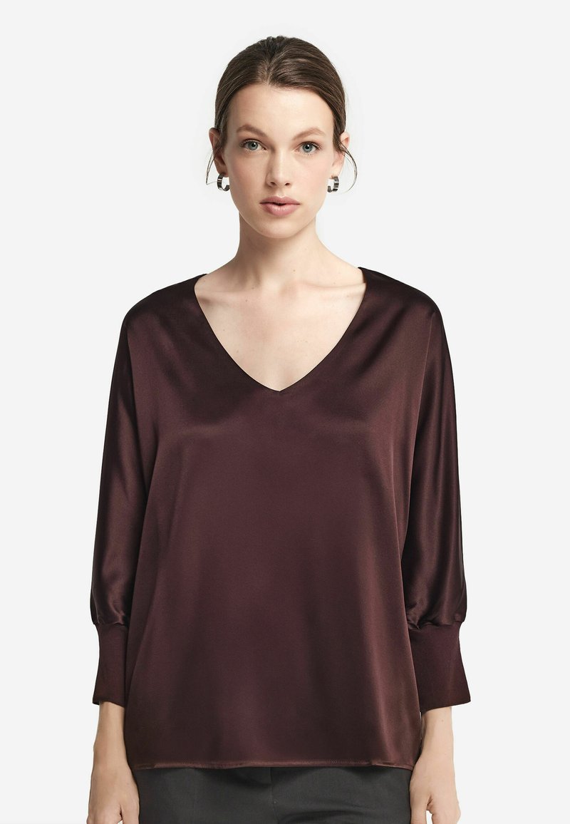 Femme portant un chemisier ample en satin bordeaux foncé à manches longues avec un col en V profond et des manches retroussées, en face sur un fond blanc.