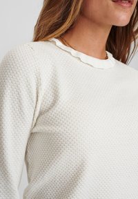 Krämfärgad tröja med ett texturerat bikake-mönster, med en volangdetalj vid halsen och mjuk, stickad tyg.