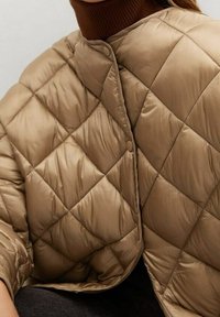 Capa acolchada beige, con cierre frontal de botones y mangas cortas, que presenta una textura suave y brillante con un patrón de diamantes.