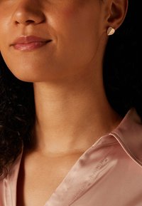 Boucles d'oreilles en forme de cœur en ton or, présentant une surface texturée, posées sur une peau lisse, avec un blouse en satin rose pâle en arrière-plan.