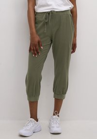 Femme portant un jogger court vert olive avec une taille à cordon et des baskets blanches, debout devant un fond clair uni.