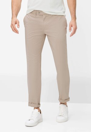 Man draagt beige slim-fit broek met omslag bij de enkels, witte lage sneakers en een wit overhemd ingestopt, staand tegen een witte achtergrond.
