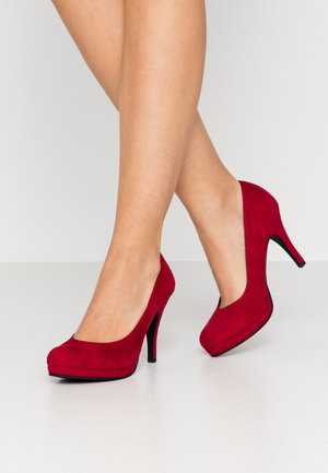 Chaussures à talons hauts en daim rouge avec un bout arrondi et une semelle noire brillante. Elles présentent une texture lisse avec un design classique de pompe.