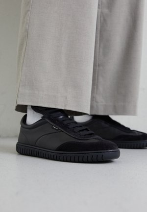Baskets basses noires avec des lacets noirs portées avec des chaussettes blanches et un pantalon beige à jambes larges.