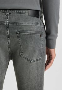 Prim-plan al unei persoane îmbrăcate în blugi gri, cu buzunar pe spate și un tricou gri cu mânecă lungă, cu mâna sprijinită lângă șold.