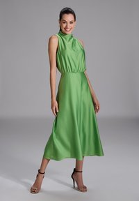 Grünes Satin-Kleid mit hohem Ausschnitt, elastischem Taillenbund und schwingendem Rock. Verfügt über ein ärmelloses Design und Knöchelriemen-Absätze.