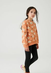 Orange blommigt sweatshirt med rund halsringning och långa ärmar, matchat med svarta leggings och kontrasterande ljusfärgade träningsskor.