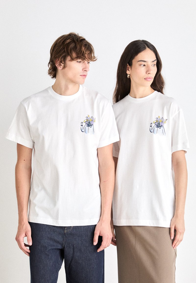 Hvide bomulds t-shirts har et lille broderet design af en figur, der holder blomster på brystet. Begge skjorter har en afslappet pasform og korteærmer.