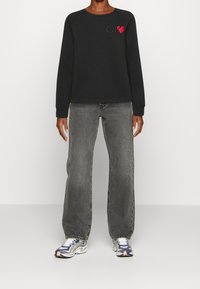 Felpa nera in cotone con scollo rotondo, caratterizzata da un piccolo cuore rosso e dal logo "CK". Abbinata a jeans larghi grigi sbiaditi e a sneaker multicolori.