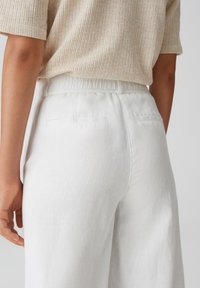 White linen trousers with a relaxed fit, featuring an elastic waistband and two back pockets. Fabric has a smooth, lightweight texture.
