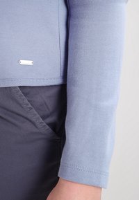 Ljusblå långärmad topp med rund hals, mjuk textur och en silverfärgad märkesetikett i fållen, tillsammans med mörkgråa tygbyxor.