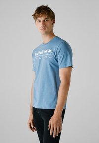 Lys blå bomulds T-shirt med rund hals, med et hvidt "Red Bull Racing" logo over brystet. Korte ærmer, tætsiddende design.