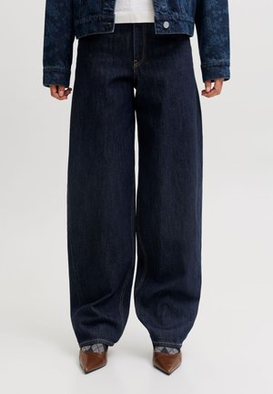 Personne portant un jean denim bleu foncé à jambes larges, des chaussures pointues marron et une veste courte en denim foncé.